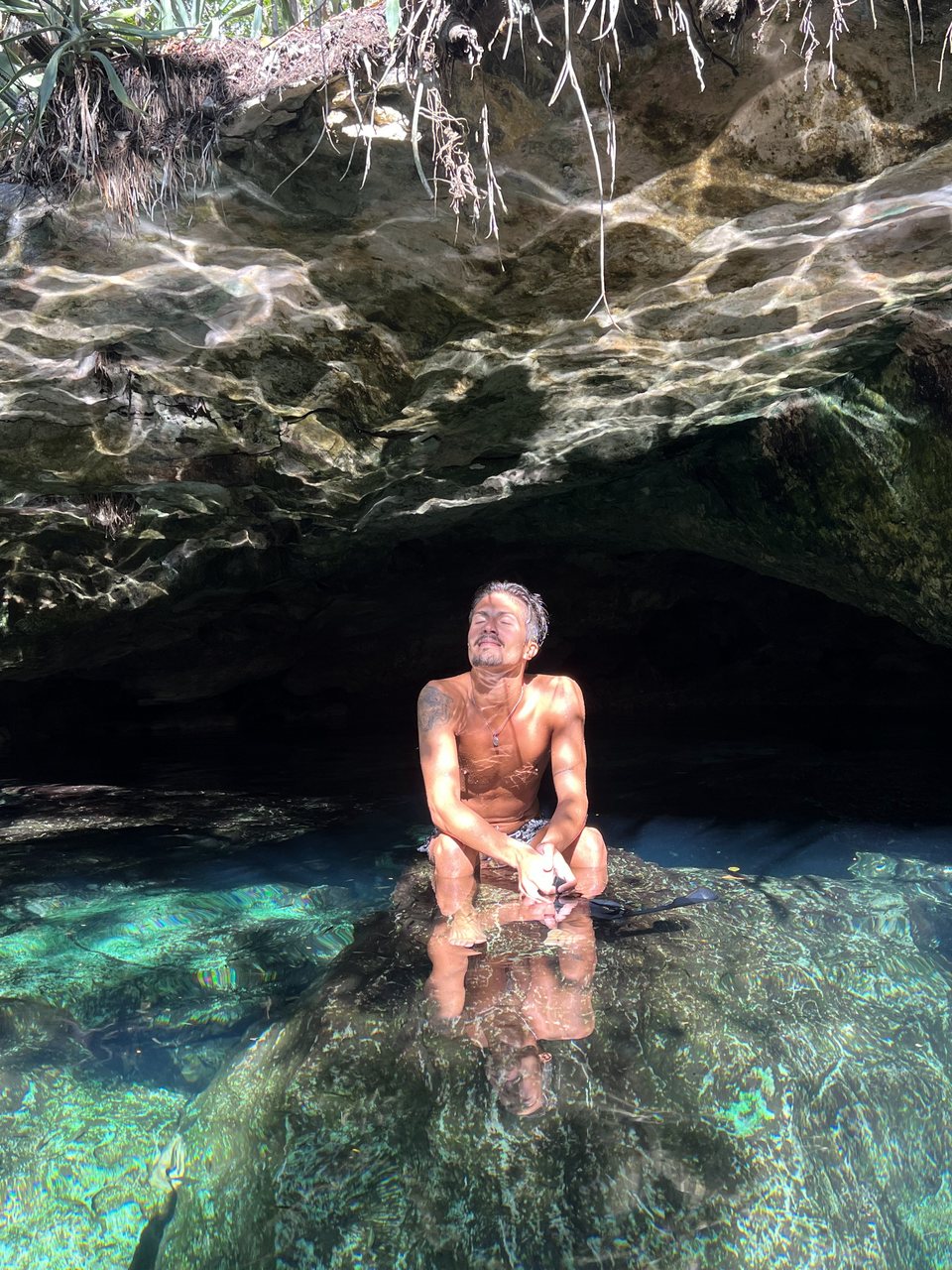 Persona sentada sobre una roca junto a un cenote, con el rostro hacia la luz del sol